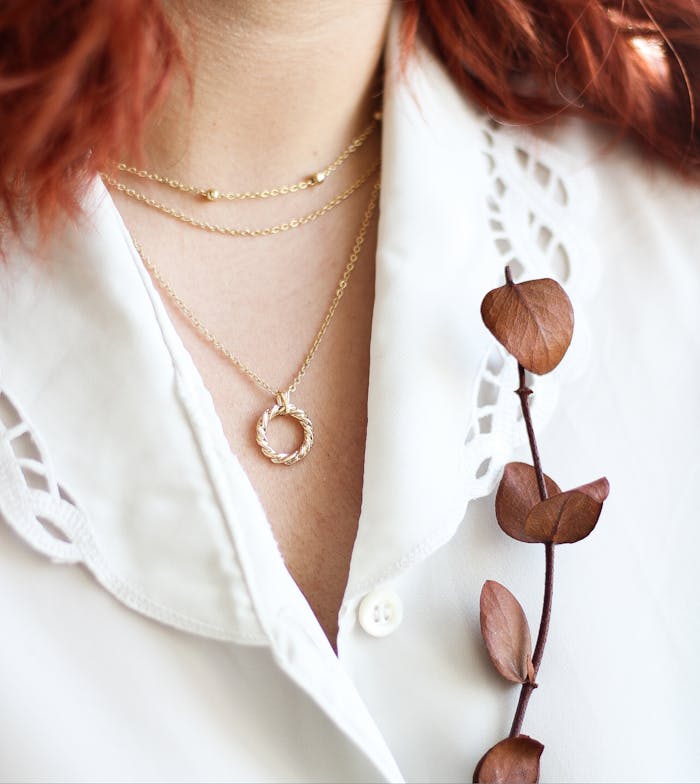 about-img Close-up of a gold necklace on a lace white shirt with red hair and dried leaves, invoking elegance.