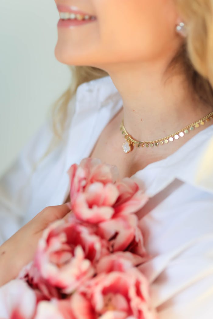 our-experience Close-up portrait of a smiling woman holding pink flowers, showcasing a gold necklace.